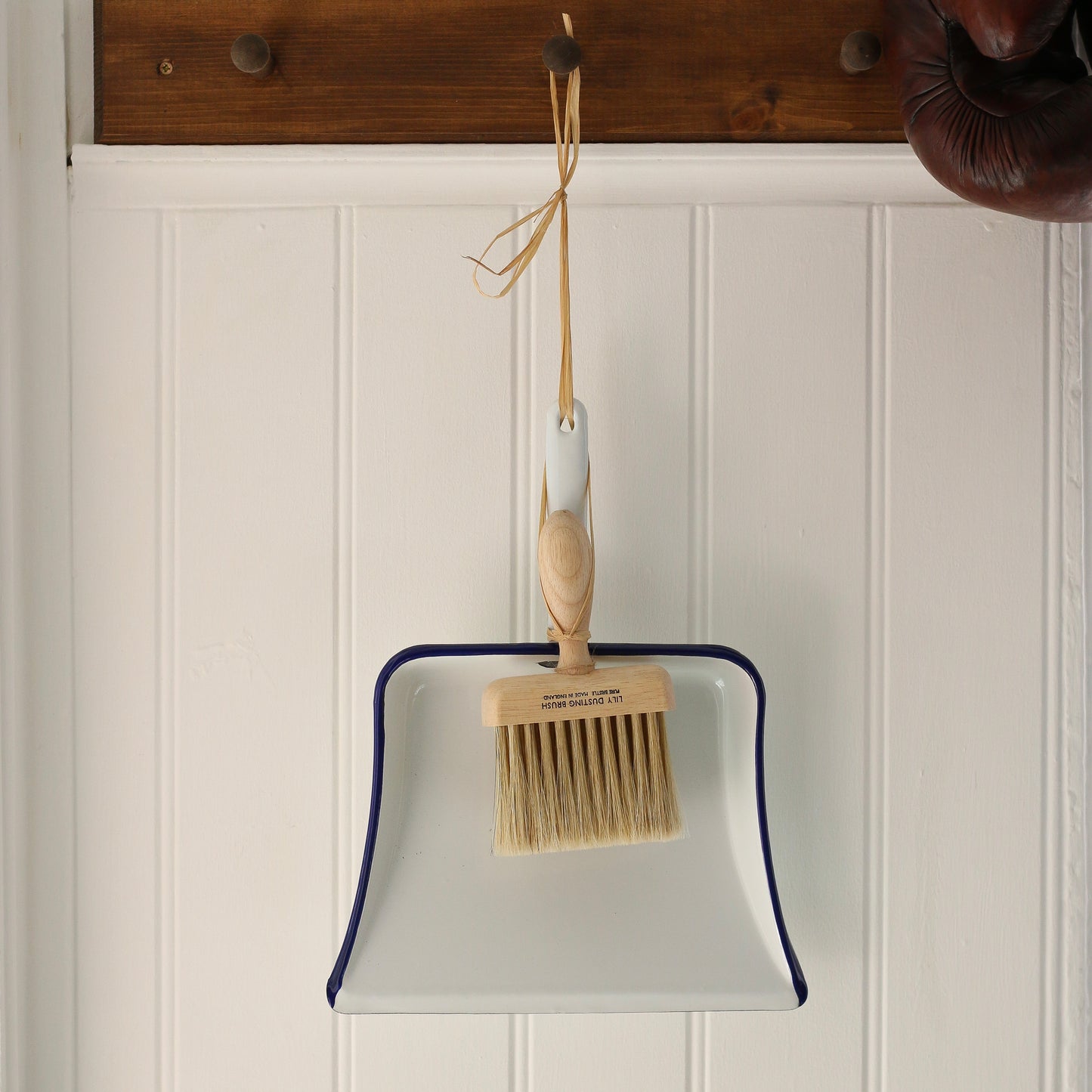 White dustpan with wooden brush hanging on a wooden hook against a white paneled wall.