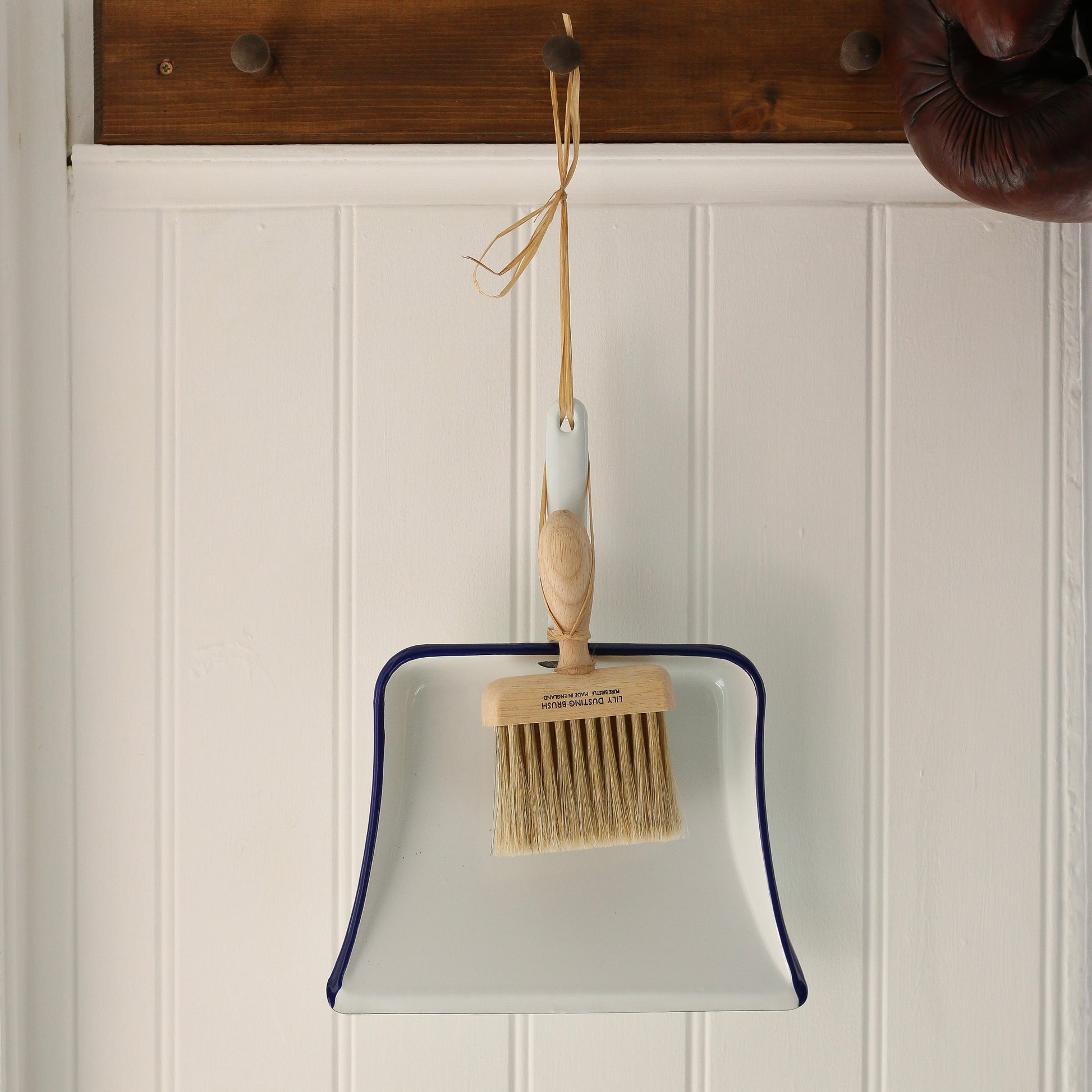 White dustpan with wooden brush hanging on a wooden hook against a white paneled wall.